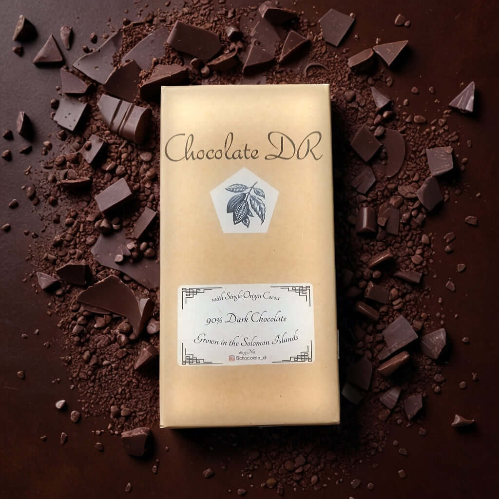 A top-down view of a chocolate bar package on a dark surface. The package is brown with a white label that reads "Chocolate DR" and "90% Dark Chocolate Grown in the Solomon Islands." Chocolate shavings and pieces are scattered around the package.