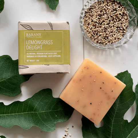 A bar of Barany Lemongrass Delight soap sits on a white surface surrounded by fig leaves and a bowl of quinoa seeds. The packaging highlights the product's natural ingredients and handmade origin in South Gippsland, Victoria.
