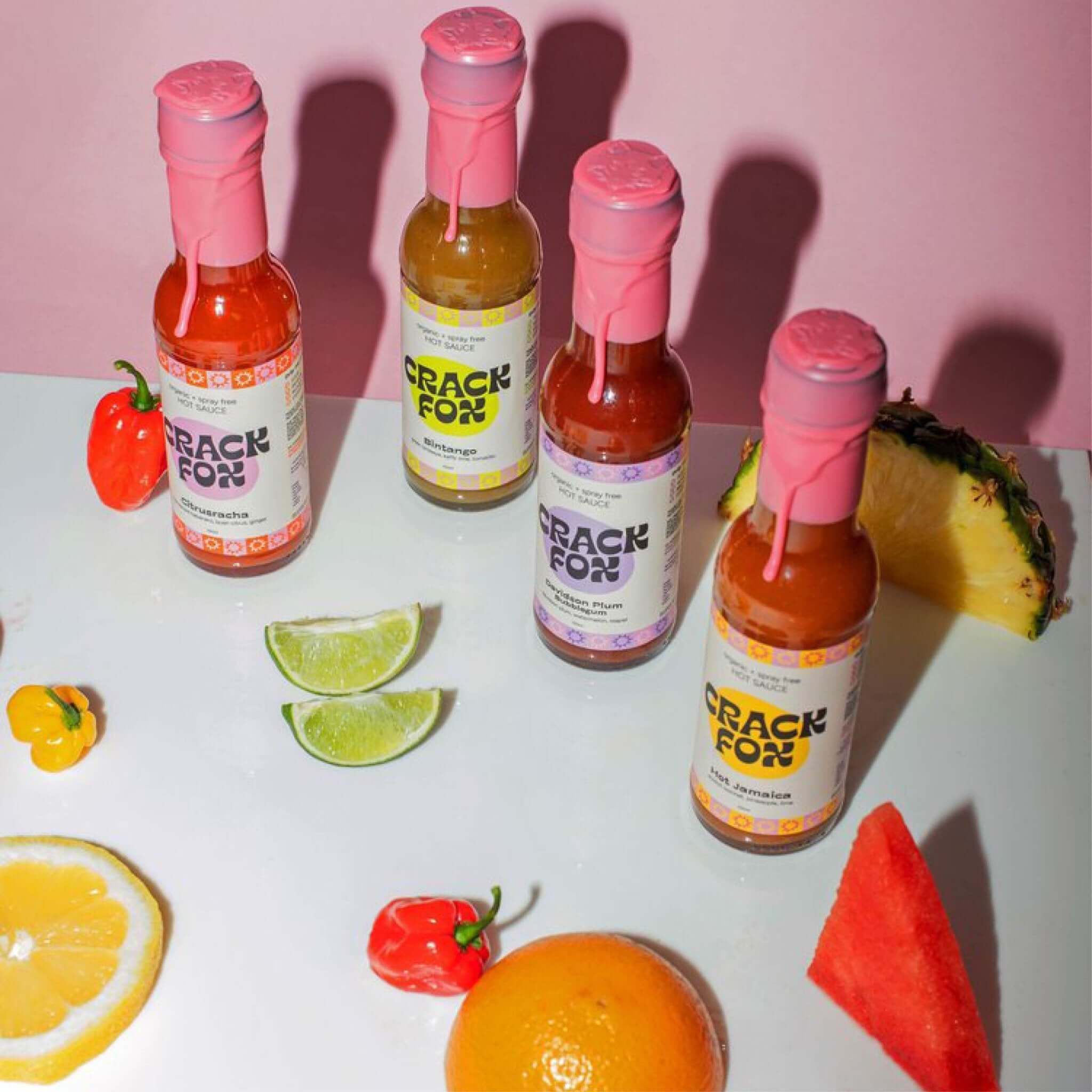 Four Crack Fox hot sauce bottles with pink wax seals, featuring flavours like Sriracha, Bintanago, Golden Plum, and Rosa Jamaica, surrounded by assorted fruits and peppers.