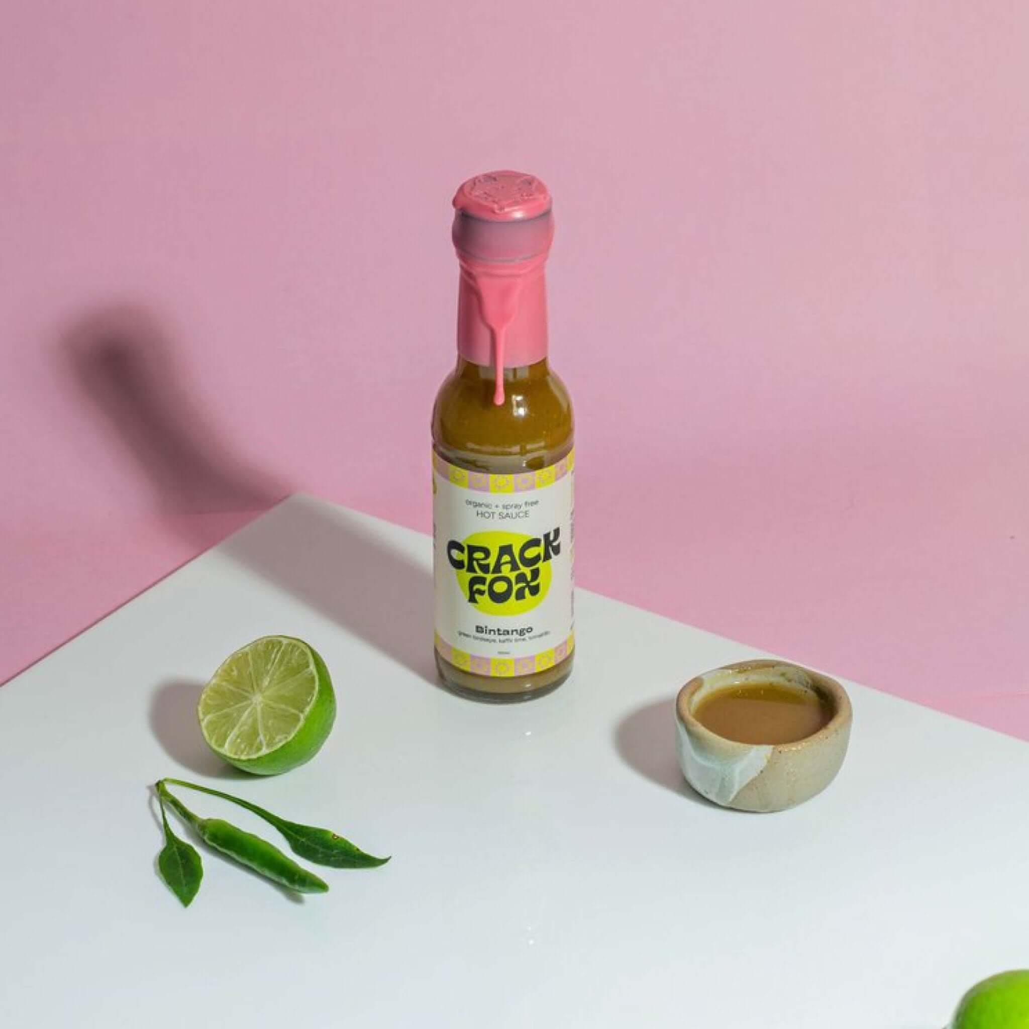 A bottle of Crack Fox Bintango hot sauce on a white surface with a pink background. Surrounded by a halved lime, green chili peppers with leaves, and a small bowl of brown sauce.