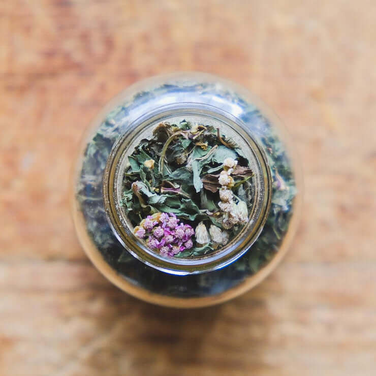 A top-down view of a glass jar filled with a colorful blend of dried herbs and flowers. The image evokes a sense of natural wellness and relaxation.