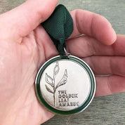 A hand holds a silver medal with a green ribbon. The medal features a stylized leaf design and the inscription "The Golden Leaf Awards."