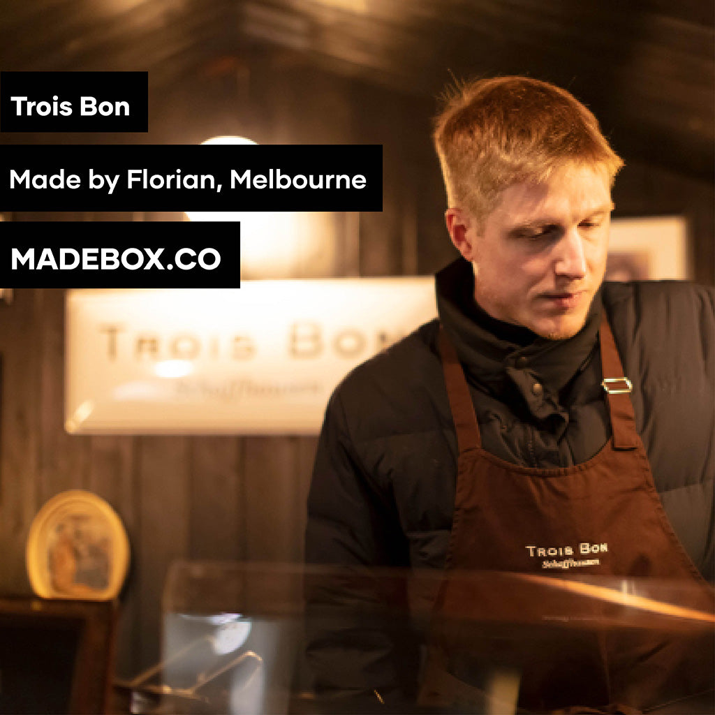  A man dressed in an apron stands behind a counter, poised to engage with patrons in a welcoming environment. The Text reads: Trois Bon Made by Florian, Melbourne