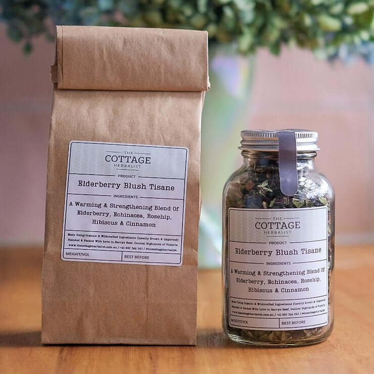 A kraft paper bag and a glass jar filled with dried herbs sit on a wooden table. Both containers feature the "The Cottage Herbalist Elderberry Blush Tisane" label and list the ingredients as a warming & strengthening blend of elderberry, echinacea, rosehi